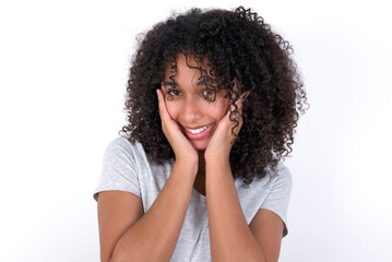 Fototapeta na wymiar Happy Young beautiful girl with afro hairstyle wearing gray t-shirt over white background touches both cheeks gently, has tender smile, shows white teeth, gazes positively straightly at camera,