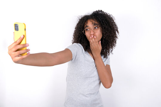 Young beautiful girl with afro hairstyle wearing gray t-shirt over white background blows air kiss at camera of smartphone and takes selfie, sends mwah via online call.