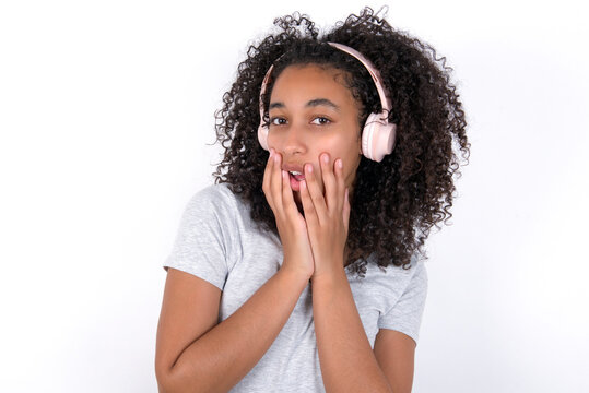 Shocked Young beautiful girl with afro hairstyle wearing gray t-shirt over white background stares fearful at camera keeps mouth widely opened wears wireless stereo headphones on ears