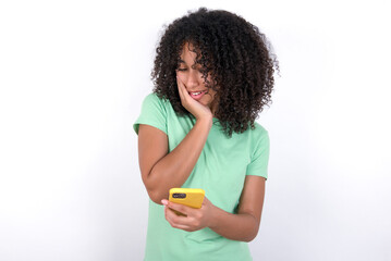 Young beautiful girl with afro hairstyle wearing green t-shirt over white background holding in hands cell reading sms using new app 5g