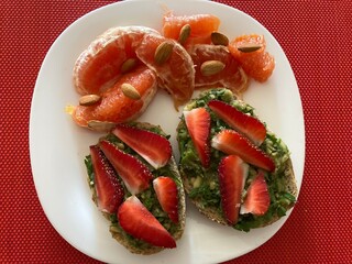 There is a white plate on a red background. On it are two toasts with greens sauce. On top are neatly sliced strawberries. Next to it are grapefruit slices and almond nuts.