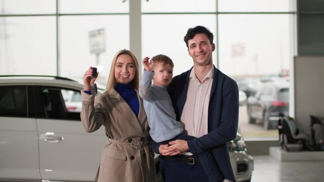 family in auto show, joyful spouses with son in his arms holding keys to new automobile while standing in auto center, smiling and looking at camera
