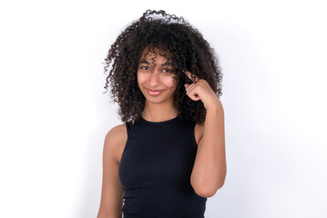 Young beautiful girl with afro hairstyle wearing black tank top over white background tries to memorize something, keeps fore finger on temple, reminds information for exam,