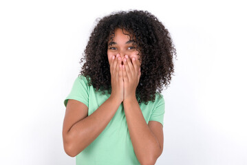 Fototapeta na wymiar Vivacious Young beautiful girl with afro hairstyle wearing green t-shirt over white background , giggles joyfully, covers mouth, has natural laughter, hears positive story or funny anecdote