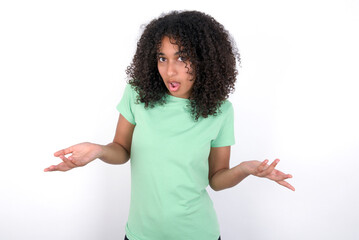 Frustrated Young beautiful girl with afro hairstyle wearing green t-shirt over white feels puzzled and hesitant, shrugs shoulders in bewilderment, keeps mouth widely opened, doesn't know what to do.