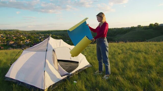 a young woman happily set up a tent on the mountain hills during sunset, for rest after a hike. a woman lays out a karimat. rest of one person in nature, in the mountains.