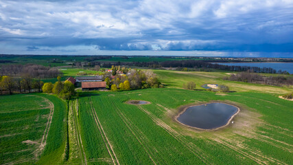 Aerial view of the village in a daylight. Green fields and small reservoir. Lake in the background. Heavy clouds in the sky.