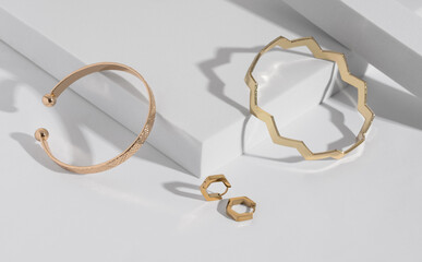 Two modern geometric shape golden bracelets and earrings pair on white podium with copy space - obrazy, fototapety, plakaty