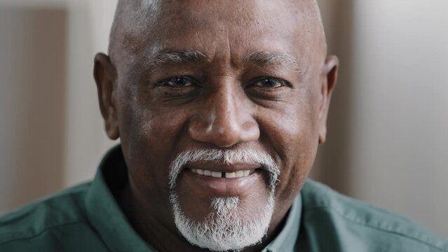 Extreme close up happy smiling old wrinkled male face. African satisfied 70s man with white healthy smile teeth in dentistry service. American elderly senior 60s grandfather looking at camera indoors
