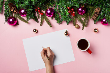 Obraz premium Notebook in the center of cute Christmas decoration: christmas balls, fir tree branch, cup coffee on pink backgraund. Female hand writing in notebook. Flat lay. Top view