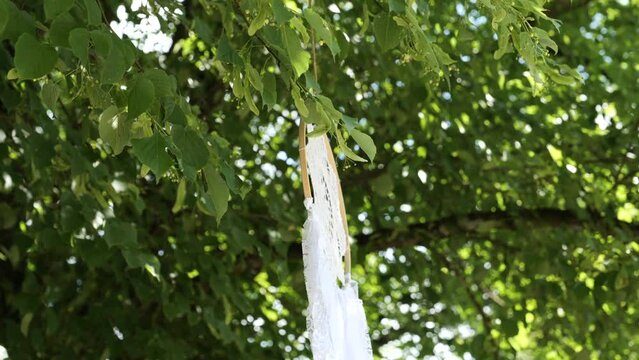 Self-made dream catcher with white fabric handing in a tree on a wedding party, slowly moving and turning in the wind. Lush, dark green leaves in the background. Tranquil, relaxing scene. 