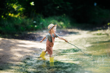 Preschooler boy in yellow rubber boots plays fishing, child fishes with stick near lake in...