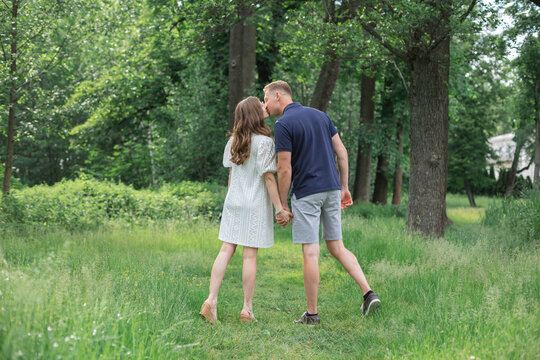 Side back view full length portrait of passionate romantic young couple kissing, holding hands in summer park