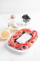 Greek salad, bowl with olive oil, bowl with olives and lemon on a white wooden background