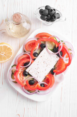 top view of greek salad, jar of olive oil, jar of olives and lemon on white wooden background