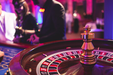 Vibrant casino table with roulette in motion, with casino chips, tokens, the hand of croupier, dollar bill money and a group of gambling rich wealthy people playing bet in the background - Powered by Adobe
