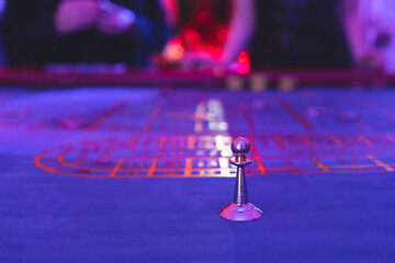 Vibrant casino table with roulette in motion, with casino chips, tokens, the hand of croupier, dollar bill money and a group of gambling rich wealthy people playing bet in the background