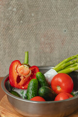 Vegetables lie in a metal bowl: tomatoes, asparagus, cucumbers, red bell peppers . on a wooden board and brown background. back gray background. place for text