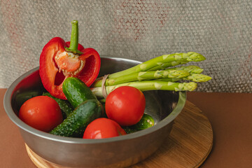 Vegetables lie in a metal bowl: tomatoes, asparagus, cucumbers, red bell peppers . on a wooden board and brown background. back gray background. place for text