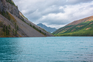 Fototapeta premium Colorful autumn landscape with big turquoise mountain lake among forest hills against sharp rock silhouette in changeable weather. Shiny ripples on azure alpine lake in sunlit valley under cloudy sky.