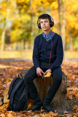 Naklejka na ściany i meble a teenage boy sits in autumn park and listens to music with headphones, trees with yellow leaves on a bright sunny day