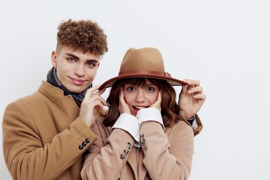 a cute couple stands on a light background in an autumn coat and sweaters. a man is standing behind her, looking tenderly at the woman, helping her to straighten her hat while she put her fingers on