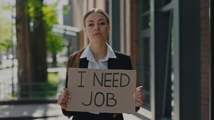 Sad laid off young woman in formal clothes standing near business center and holding cardboard placard with I need job phrase. Unemployment growth concept.