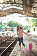 Young beautiful woman of 30-40 years old with a pink small suitcase is waiting for her train at the station. Curly model with casual clothes talking on the phone, drinking coffee to go. Travel.