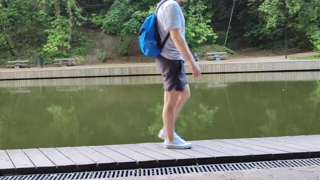 Young white caucasian man walks along the shore of a pond. Man in sports clothes and with a backpack on his back is walking slowly along the pier. Walk near the lake or river in the city park