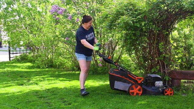 Woman mowing green lawn in backyard with lawn mower and leaning on bench on summer sunny day, slow motion. Landlady cleaning up territory, blooming bushes background. Maintenance of home garden.