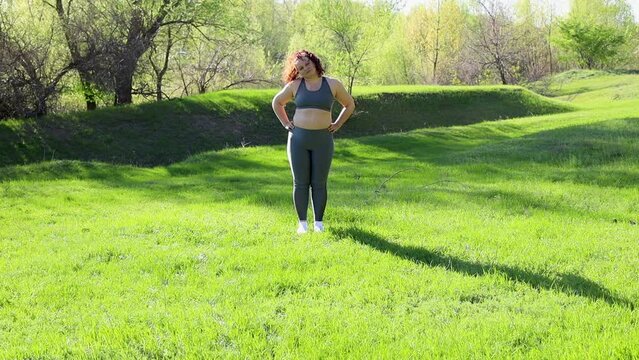 Young overweight woman in fitness suit tilt head from side to side standing on green lawn in park on sunny day, nature background. Healthy lifestyle, exercise and fitness outdoors. Sports activity.