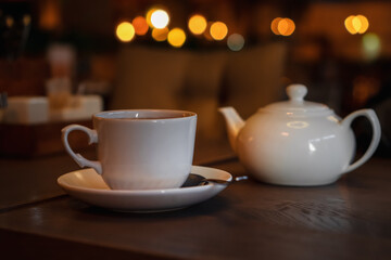 Fototapeta na wymiar White cup with teapot. Pot standing on saucer in soft focus on naturally blurred background. Coffee, tea house, bokeh lights. The concept of a cozy pastime, tea ceremony