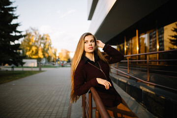 Attractive stylish caucasian long-haired model with big lips in a warm burgundy jacket poses while leaning on iron railings in the city, autumn evening.