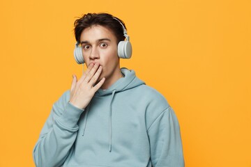 a happy man in a blue hoodie stands on an orange background listening to music with headphones covering his mouth with his hand in surprise