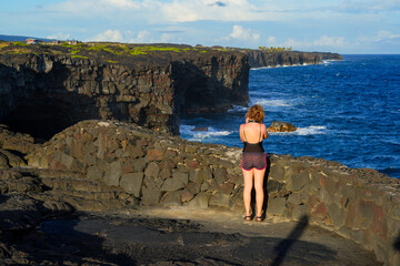 Blonde girl taking some pictures of the cliffs of lava rock plummeting into the Pacific Ocean located on the southern side of the Hawaiian Volcanoes National Park on the Big Island of Hawai'i