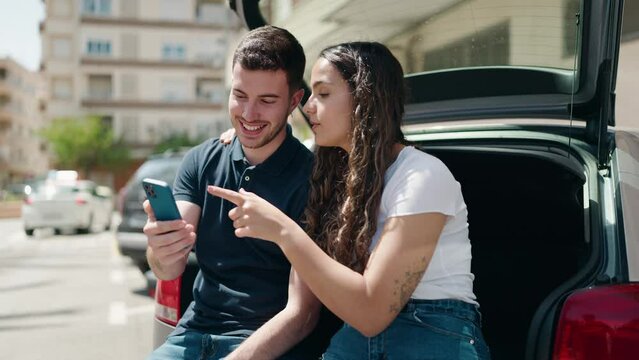 Young hispanic couple sitting on car trunk using smartphone at street