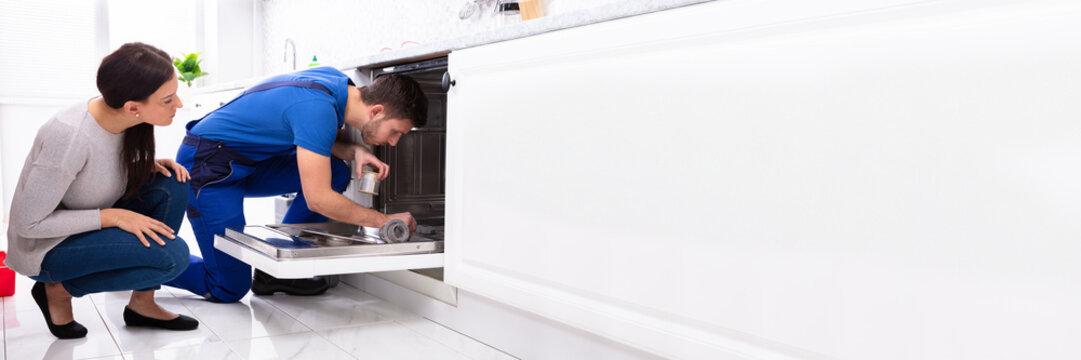 Woman Looking At Repairman Repairing Dishwasher In Kitchen