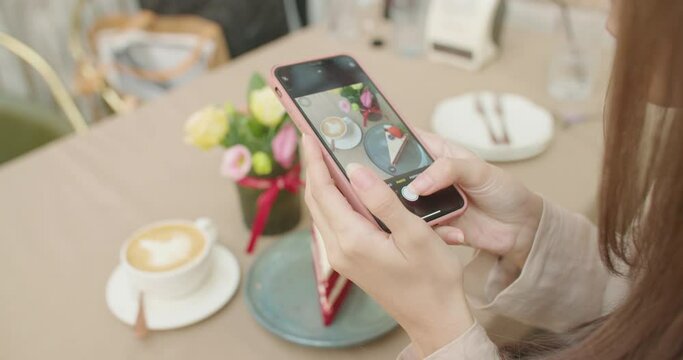 Close up of woman holds smartphone taking pictures of sweet dessert and coffee to share photos on social media. Attractive satisfied young girl feeling pleasure with delicious food cake at coffeeshop.