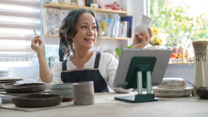 Smiling senior female holding pottery earthenware product in front of digital tablet while live streaming. E-commerce, online selling.