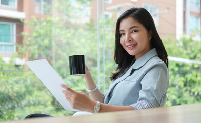 Confident young businesswoman drinking coffee and reading document at modern workplace in the morning.