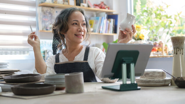 Smiling senior female holding pottery earthenware product in front of digital tablet while live streaming. E-commerce, online selling.