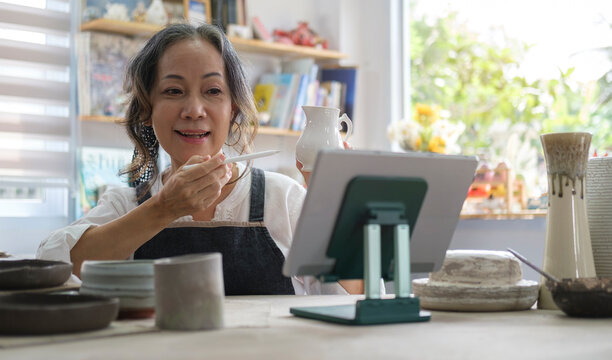 Mature woman holding pottery earthenware product in front of digital tablet while live streaming. E-commerce, online selling, small business concept.