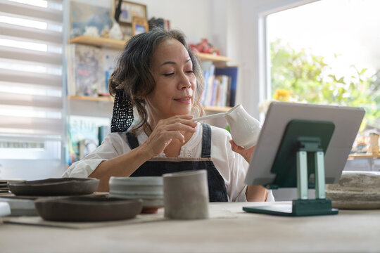 Small ceramic studio owner holding pottery earthenware product in front of digital tablet while live streaming on internet for selling.