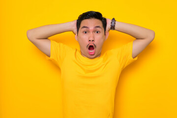 Shocked young Asian man in casual t-shirt with open mouth looking at camera in amazement isolated on yellow background. People lifestyle concept