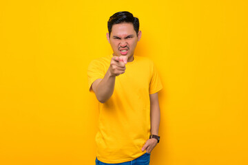 Angry young Asian man in casual t-shirt pointing finger at camera isolated on yellow background. People lifestyle concept