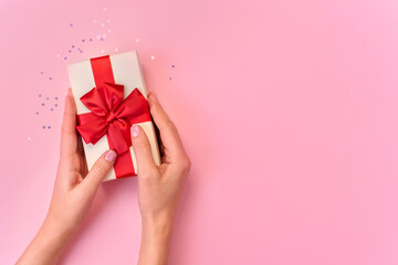 Hands of young girl are holding gift box with red bow on pink background with confetti. Top view.