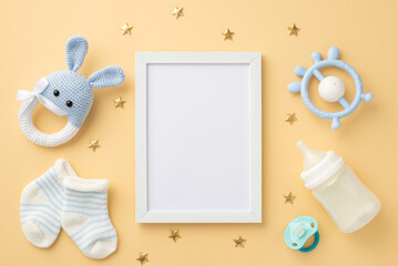 Baby accessories concept. Top view photo of photo frame knitted bunny rattle toy blue teether milk bottle tiny socks baby's dummy and gold stars on isolated pastel beige background with blank space