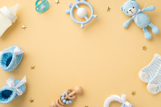 Baby concept. Top view photo of blue teddy-bear toy rattle socks milk bottle teether soother knitted shoes and gold stars on isolated pastel beige background with empty space in the middle