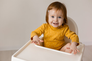 funny red-haired blue-eyed baby in a yellow bodysuit sitting in a white high chair