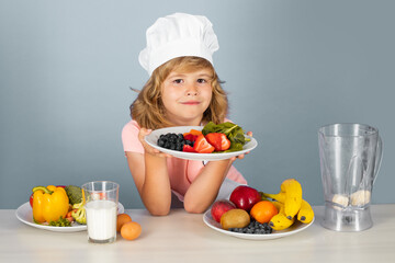 Child chef hold plate with fruits isolated on blue. Funny little kid chef cook wearing uniform cook cap and apron cooked food in the kitchen.